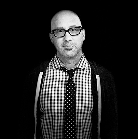 "A black and white image of man with glasses wearing a checkered shirt and a tie"