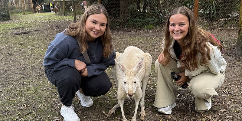 Two girls kneel down by a kangaroo within a forest.