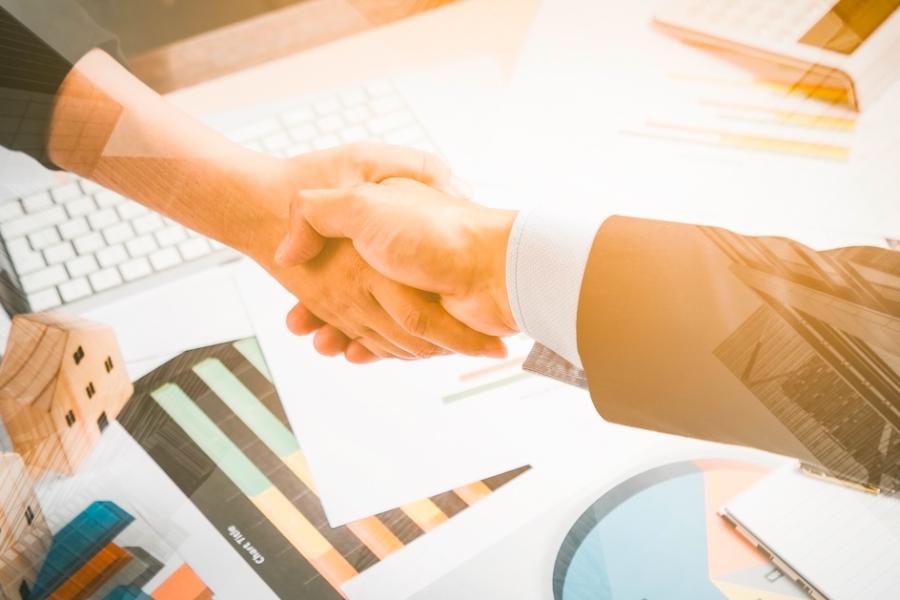 Two unseen people shake hands over a desk littered with business tech and materials.