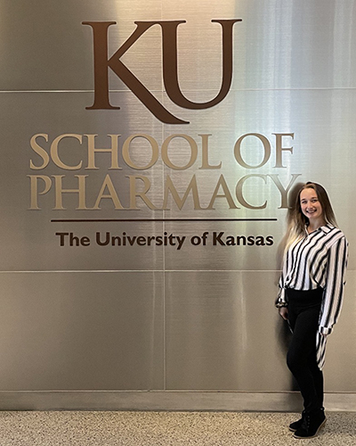 A person stands smiling in front of a wall with the words "KU School of Pharmacy, The University of Kansas" displayed.