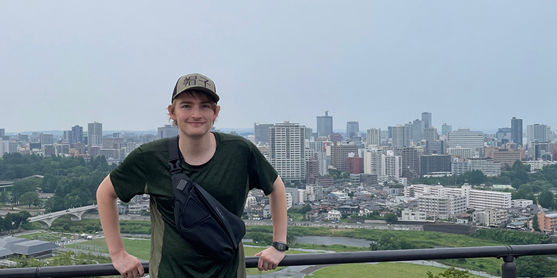 A person wearing a cap and shirt stands smiling at a scenic viewpoint overlooking the city of Tokyo. Tall buildings and urban landscape extend into the distance under a clear sky.