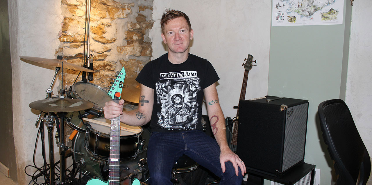 Brad Osborn in his home studio, seated on drum stool, holding guitar in right hand.