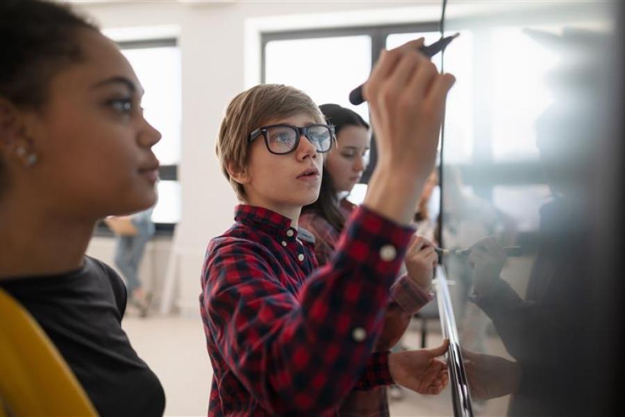 A group of students in front of a whiteboard. One of the students is holding a marker and writing on the board.