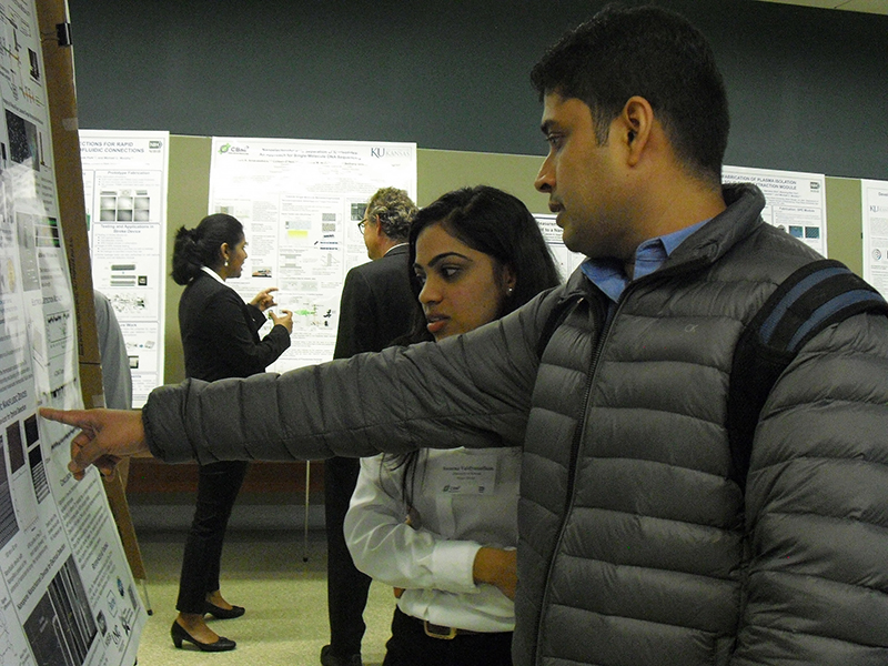 Two people in front of a research poster, one pointing to specific details.