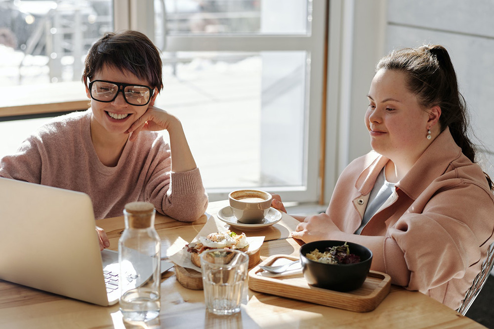 A woman wearing glasses sits next to a woman with Down Syndrome as they both look at a laptop computer over breakfast. 