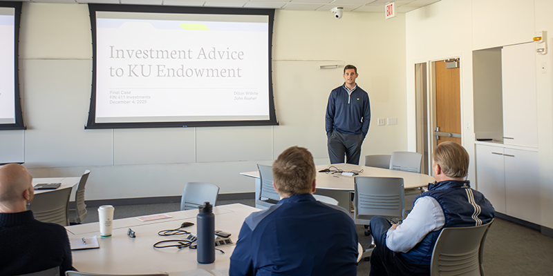 A person stands at the front of a classroom presenting a slideshow titled "Investment Advice to KU Endowment." Several people are seated around a table, facing the presenter.
