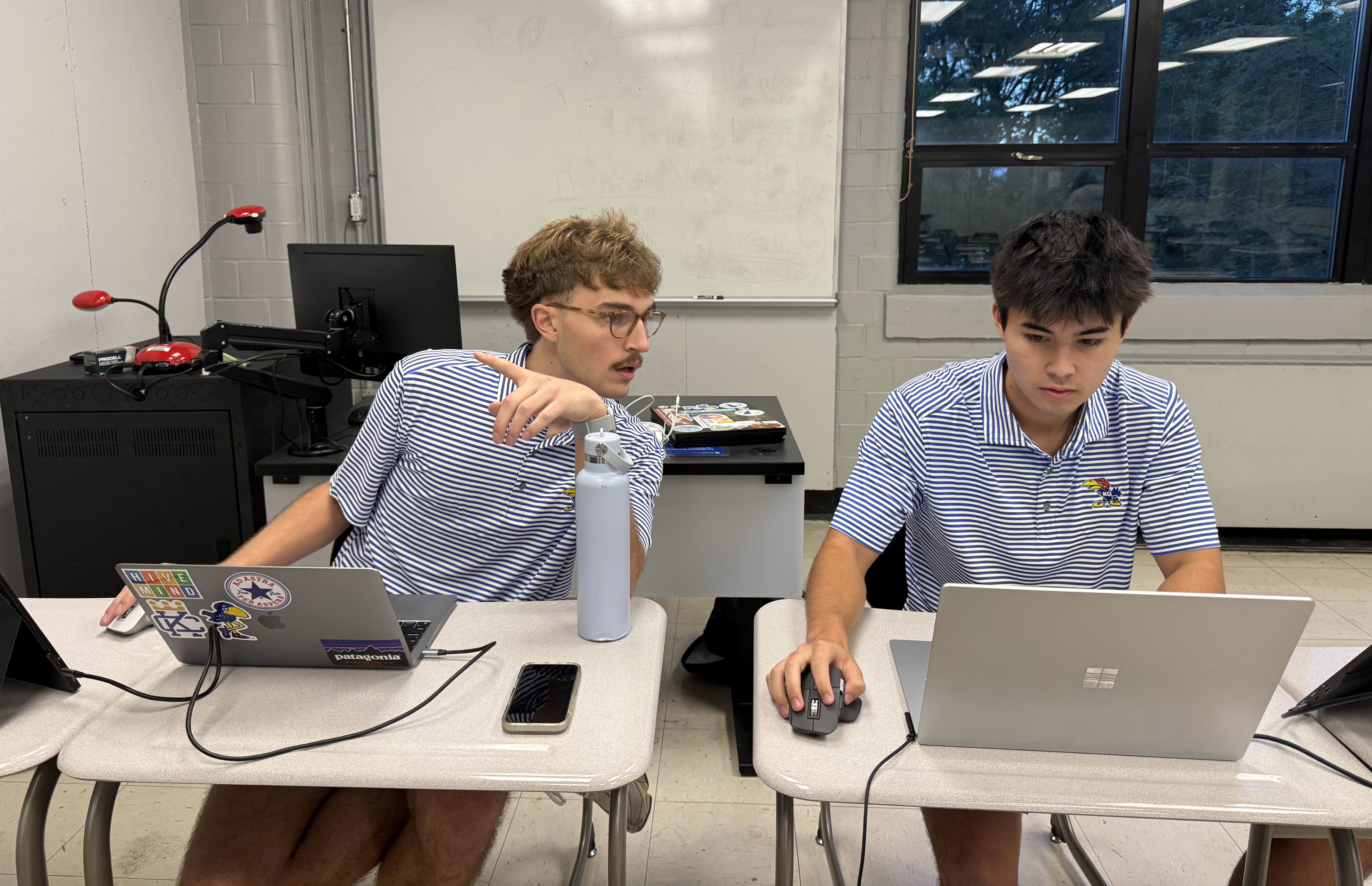 Owen Williams and Zach Willingham work at computers on desks in classrooms at debate tournament.
