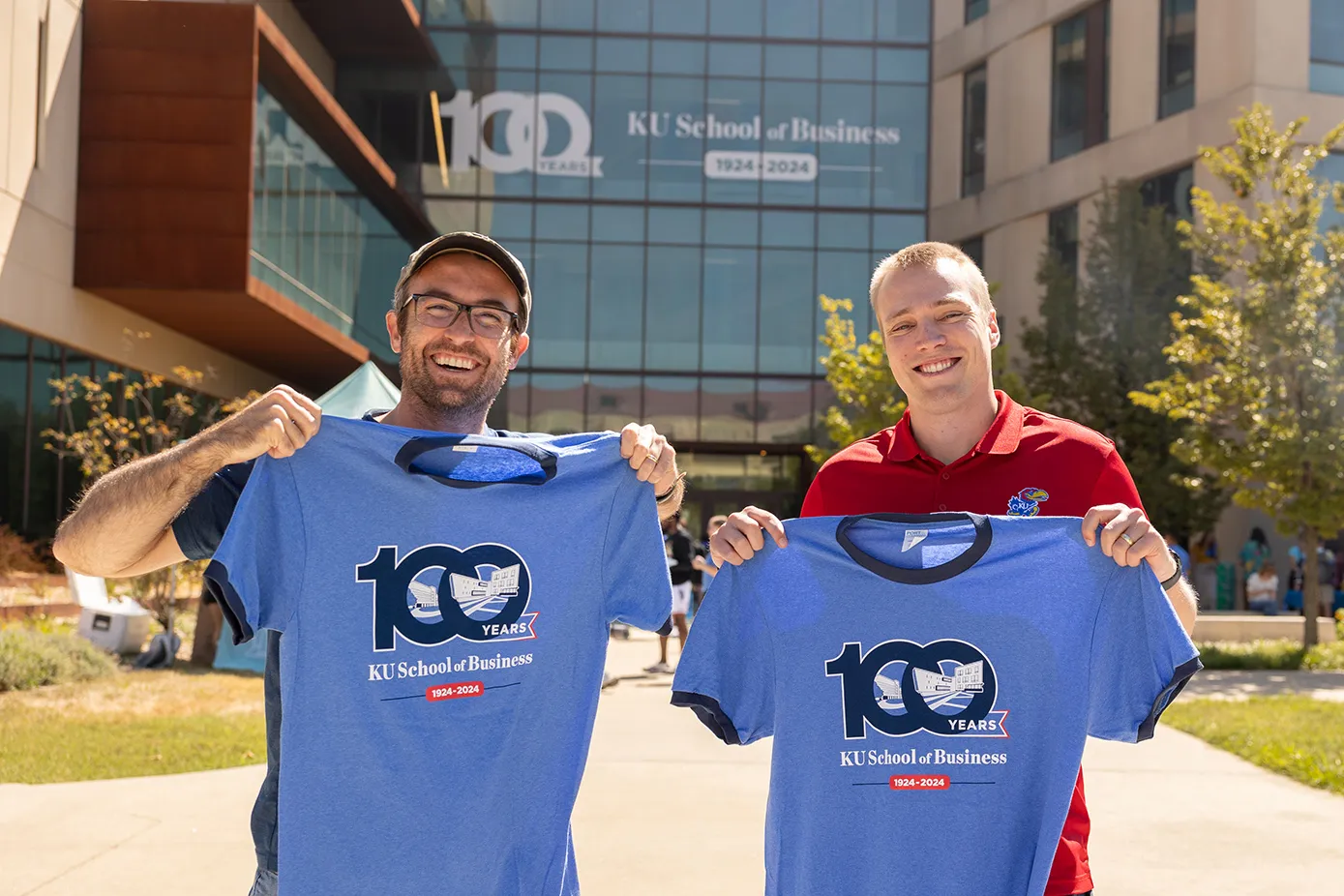 Two individuals proudly displaying commemorative t-shirts for the 100th anniversary of KU School of Business in front of Capitol Federal Hall.
