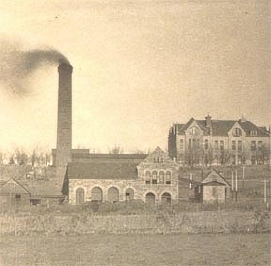 Historic photo of the original KU Powerhouse with a tall smokestack emitting smoke, showing nearby campus buildings in the background.