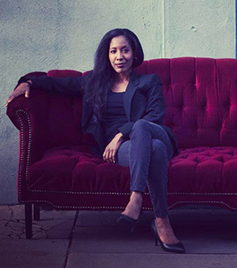 photo of a woman sitting with her legs crossed on a couch. Her arm is slung over the arm of the couch.  She looks directly at the camera