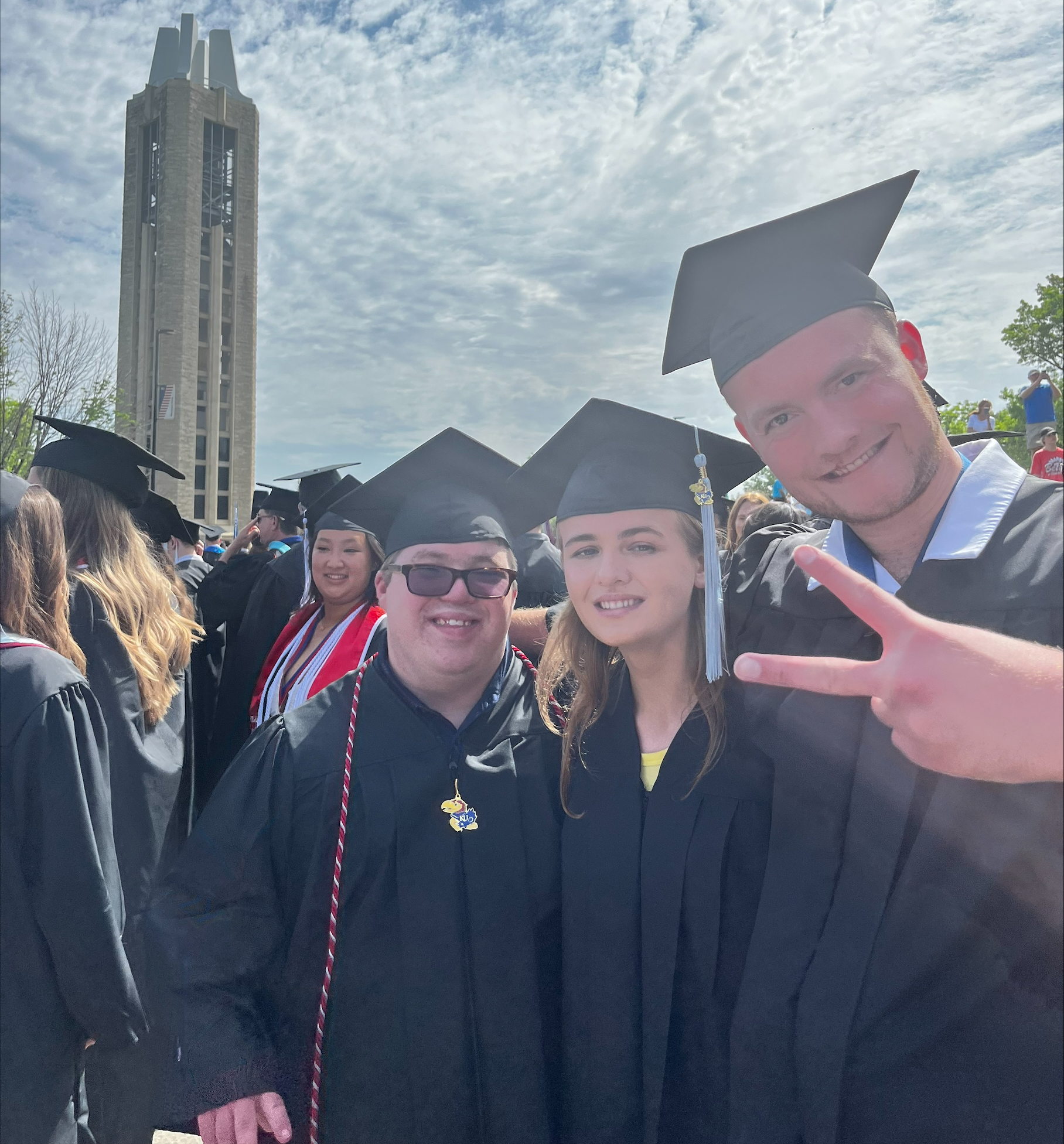 Students on graduation day wearing caps and gowns and looking at the camera. The background is the KU Campanile and it's a sunny day.