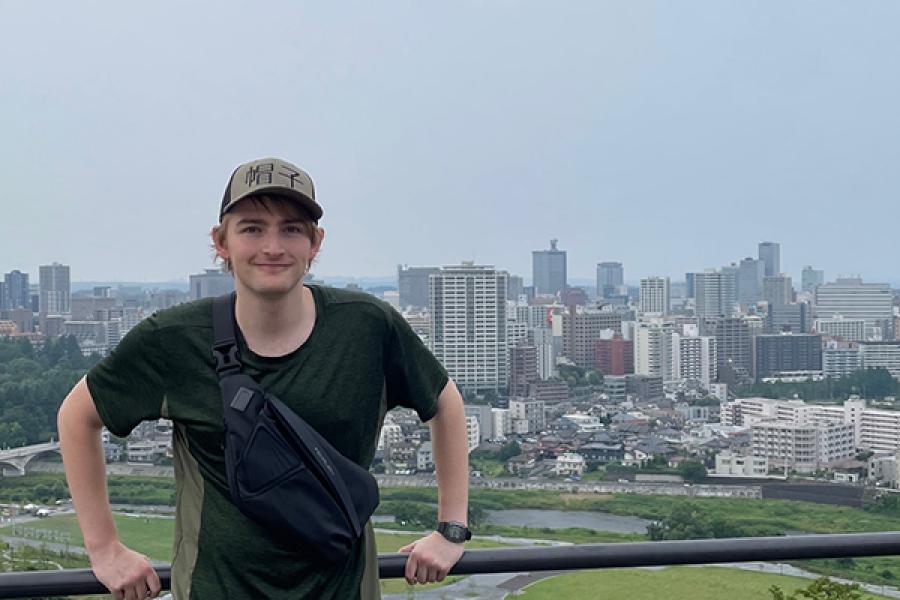 A person wearing a cap and shirt stands smiling at a scenic viewpoint overlooking the city of Tokyo. Tall buildings and urban landscape extend into the distance under a clear sky.