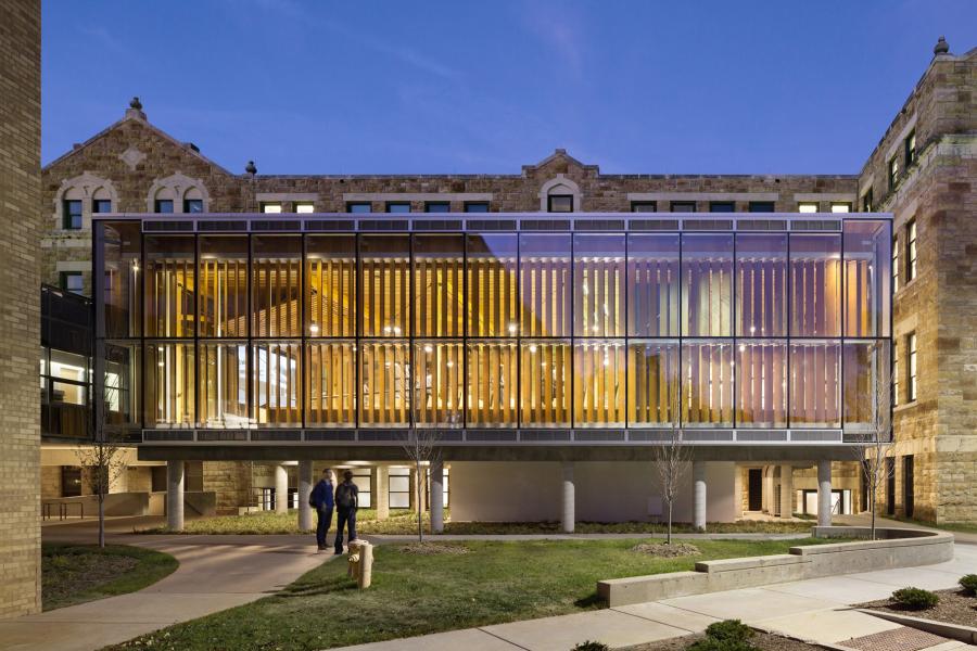 Exterior view of the glass and wood facade of the Forum at Marvin Hall at the University of Kansas during twilight, with two individuals walking in front.