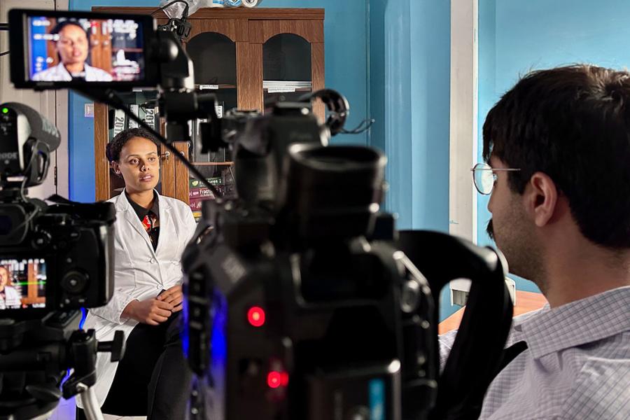 Man standing at right, operating a camera, interviewing woman seated at left.