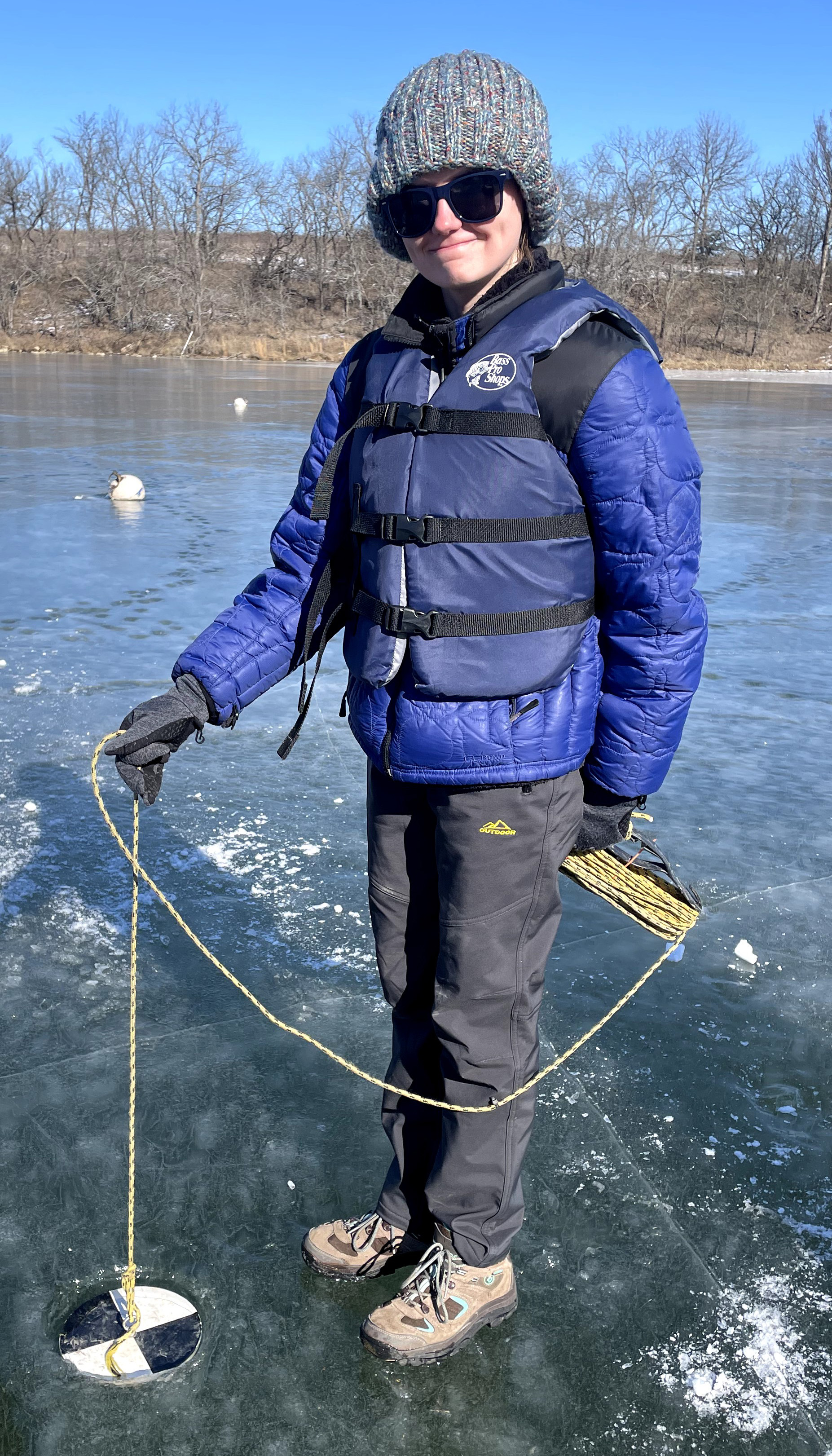 Alexandra Coveney standing on a frozen pond lowering a black-and-white Secchi disk device, attached to a rope, through a hole into the water.