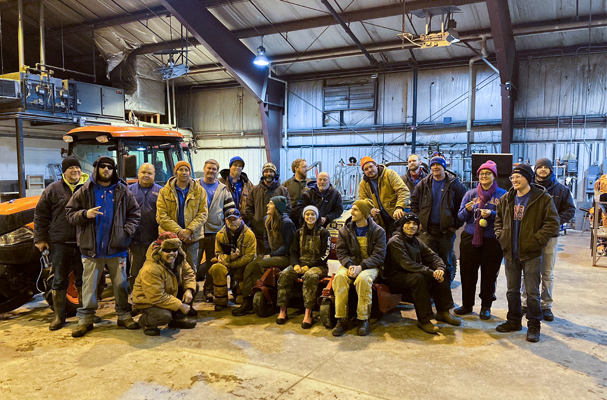 Standing and seated in a campus facility, wearing heavy winter clothing, the KU grounds crew poses for a group photo after a morning of snow and ice removal.