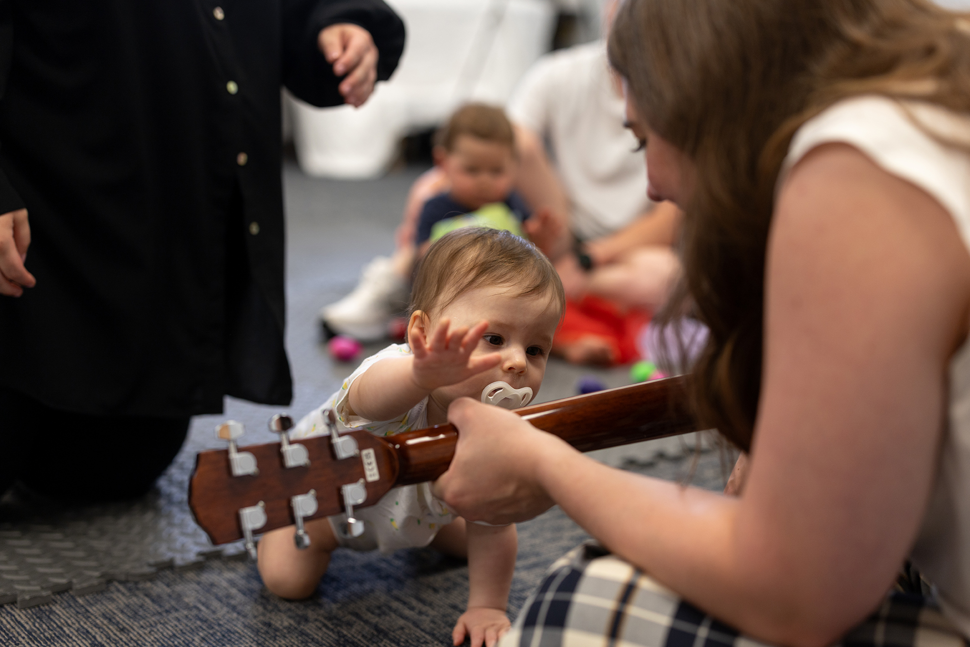 A baby sucking a pacifier craws on the floor and reaches for a guitar being played by a woman sitting on the floor