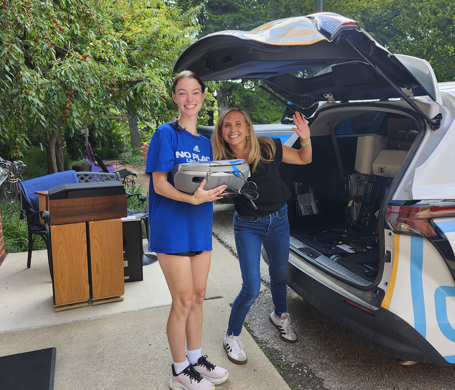 A tall young woman wearing a blue KU t-shirt holds a gray boom box and prepares to hand it to a blond woman who waves at the camera. They are both next to an open van door with speakers and other audio equipment in the background.