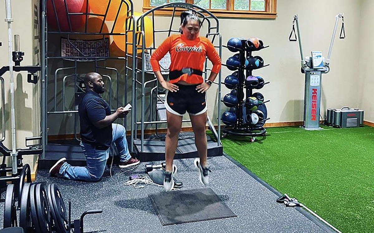 An athlete jumps with a band around her waist to measure height and force, while a researcher looks on.