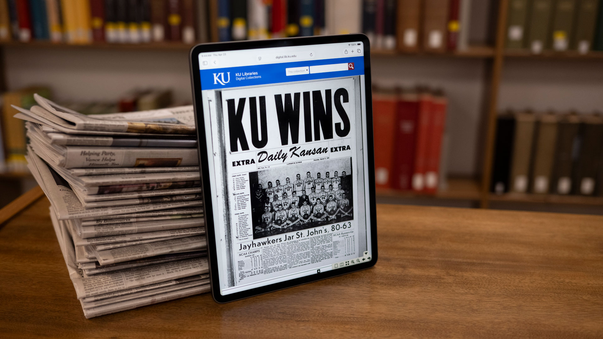 An ipad leaned up against a stack of newspapers displays a digitized Daily Kansan telling of the 1952 basketball national championship.
