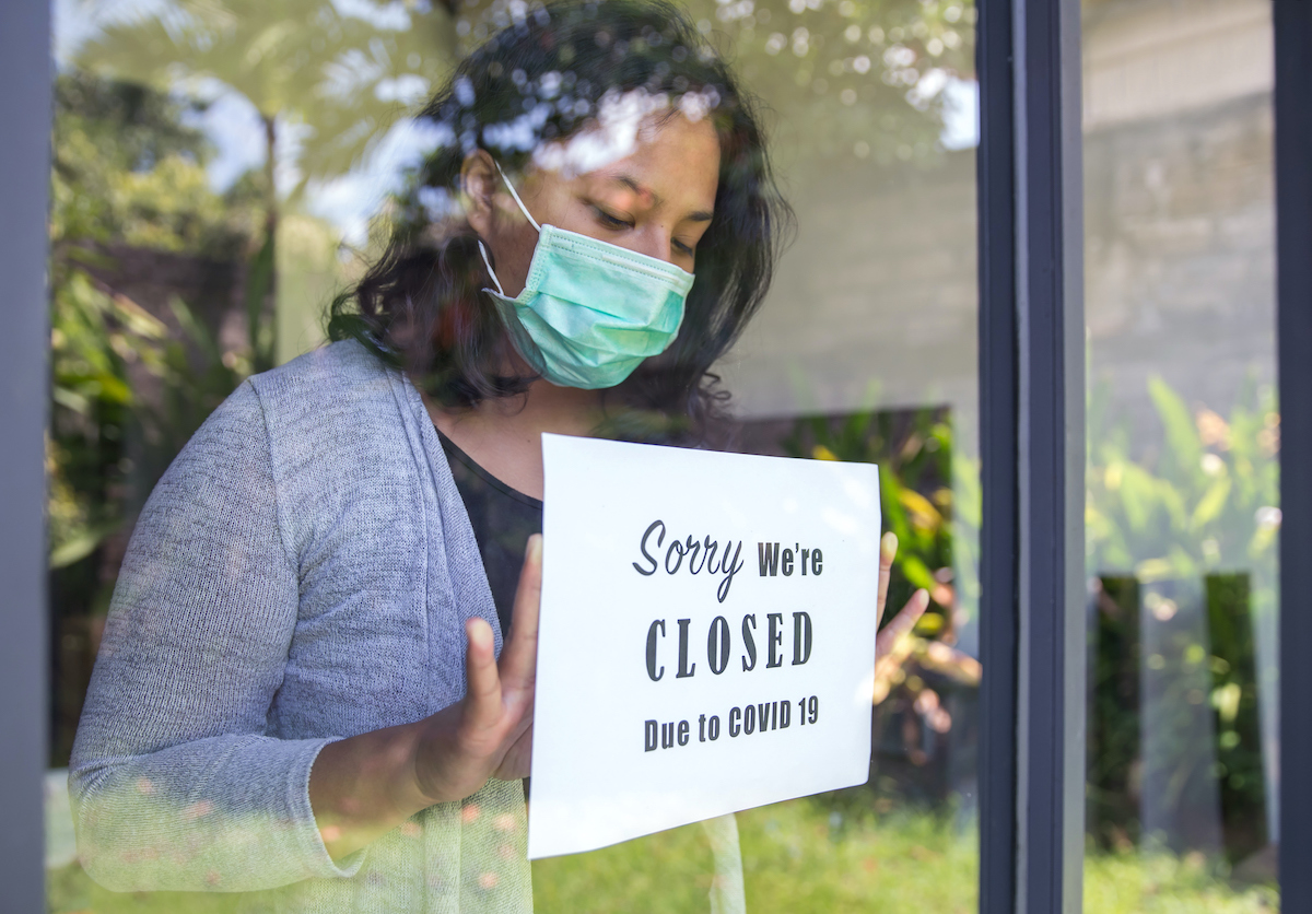A woman wearing a medical mask puts a sign in a window front with the text "sorry we're closed due to COVID-19"