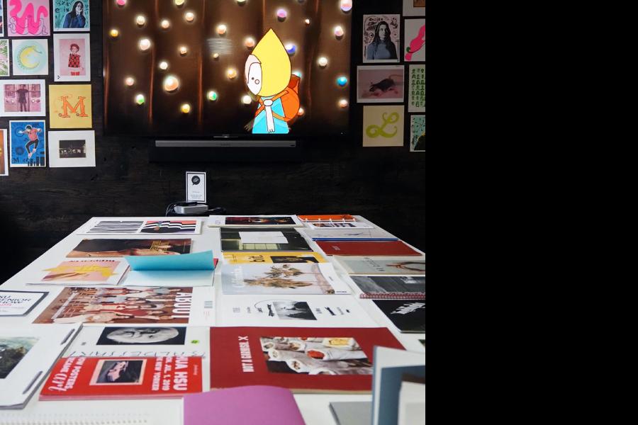 Gallery workspace with a table covered in printed design work and a wall mounted screen displaying an animated character, surrounded by framed posters.