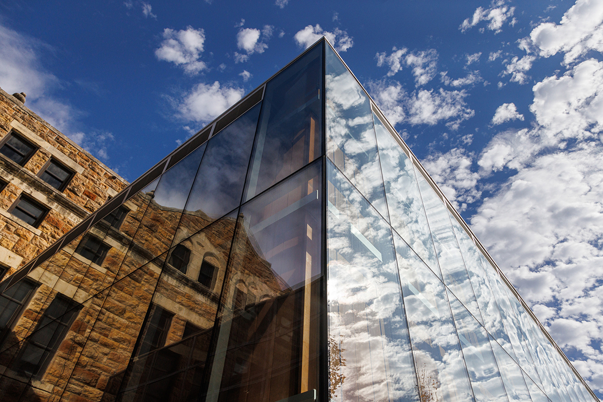 Reflective glass building corner juxtaposed against an older stone structure under a blue sky with scattered clouds.