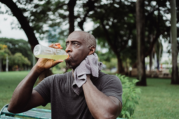 An adult man with brown skin and graying hair is mopping sweat from his neck and drinking a light green beverage.