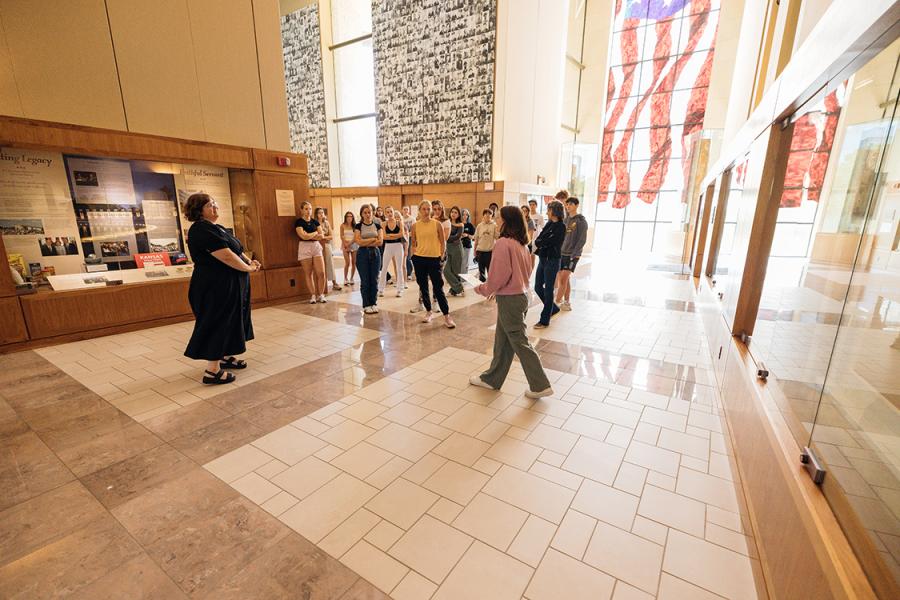 Visitors gather in the main hall of the Dole Institute of Politics at the University of Kansas. A stained-glass flag and solider memorial memory wall are visible in the background.