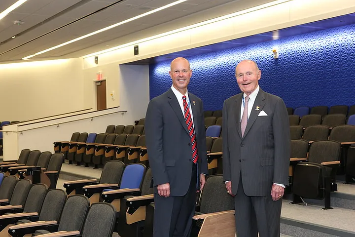 Two men standing in an auditorium with rows of seats and a blue, textured wall in the background.