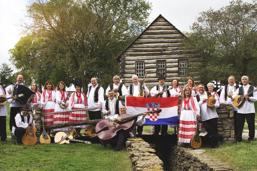 Outdoor group photo of Hrvatski Običaj, a 20-piece Croatian band, in front of a log cabin.