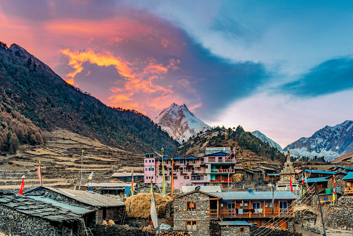 Village in Nepal at base of mountains with pastel sky, sun behind clouds.