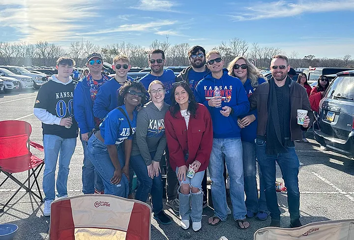 Group of people at a tailgate party in a parking lot, wearing Kansas City and Kansas Jayhawks sports apparel, smiling and holding drinks under a clear sky.