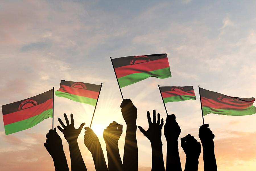 A photo of several young people's hands holding flags of Malawi in the air, with the sun in the background.