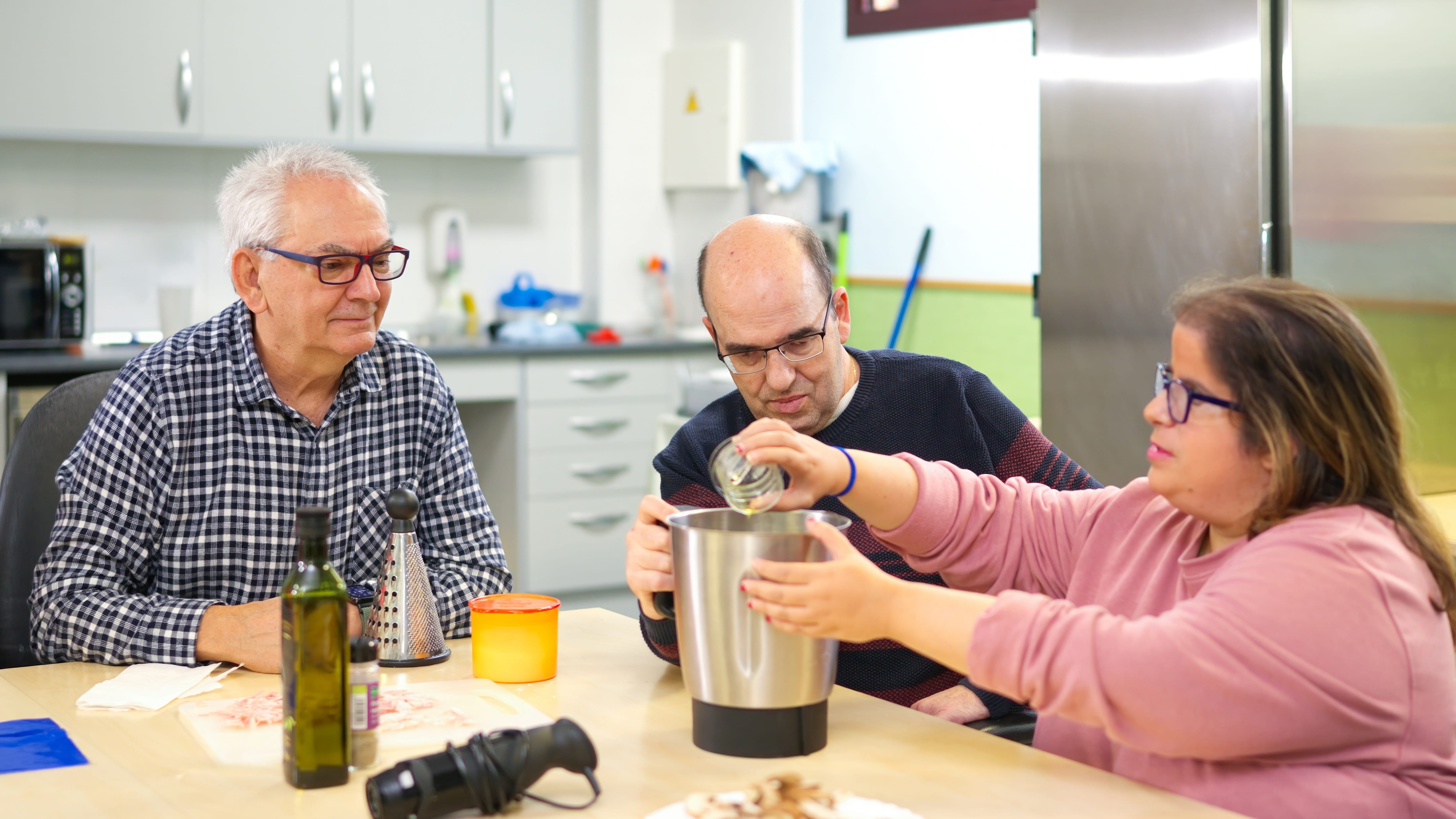 Three adults sit at a table and combine ingredients while cooking dinner in their home.
