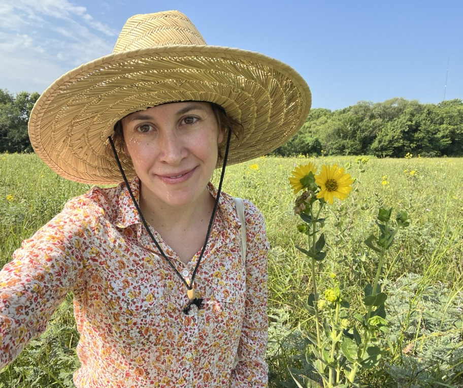 Young person wearing straw hat and floral shirt, standing in a field looking at camera.