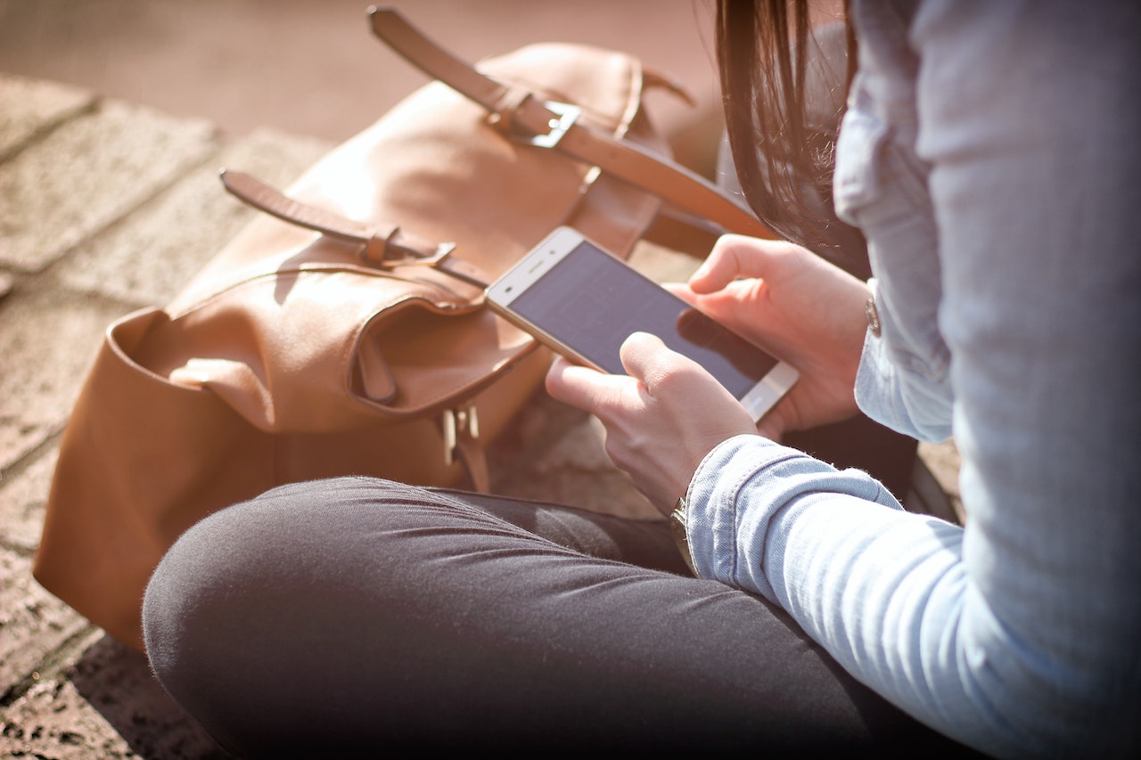 "A woman holds a phone while sitting  on a brick surface next to a large bag"