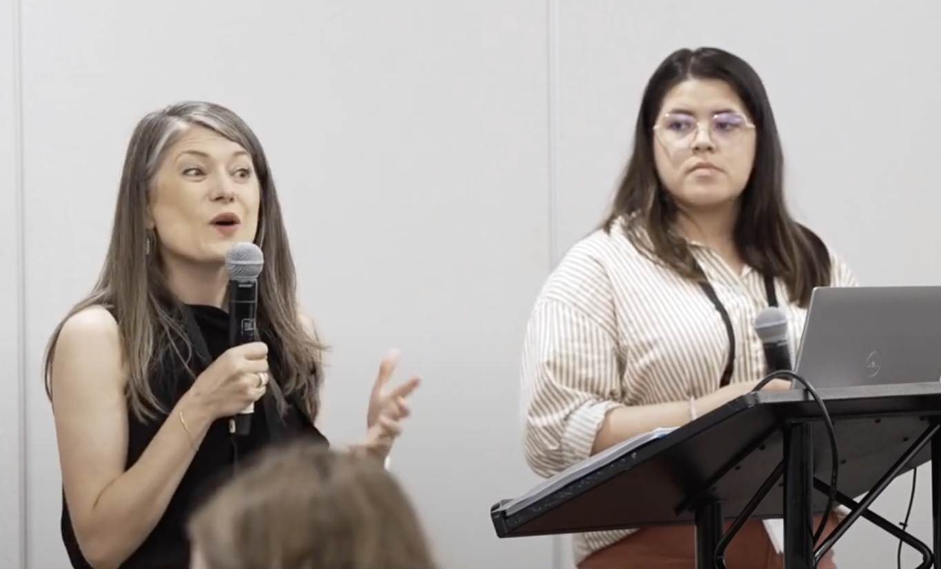 A woman with long hair speaks into a microphone from behind a podium while next to another woman with long hair and glasses also holding a microphone