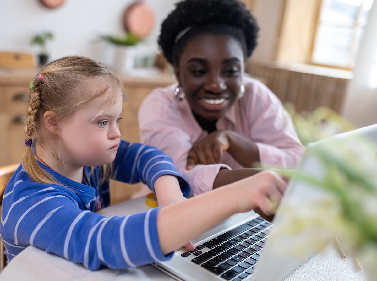 "A student with braided blonde hair wearing a blue striped shirt who has Down Syndrome points to a computer screen while a teacher with short, curly black hair and a pink blouse looks at her work."