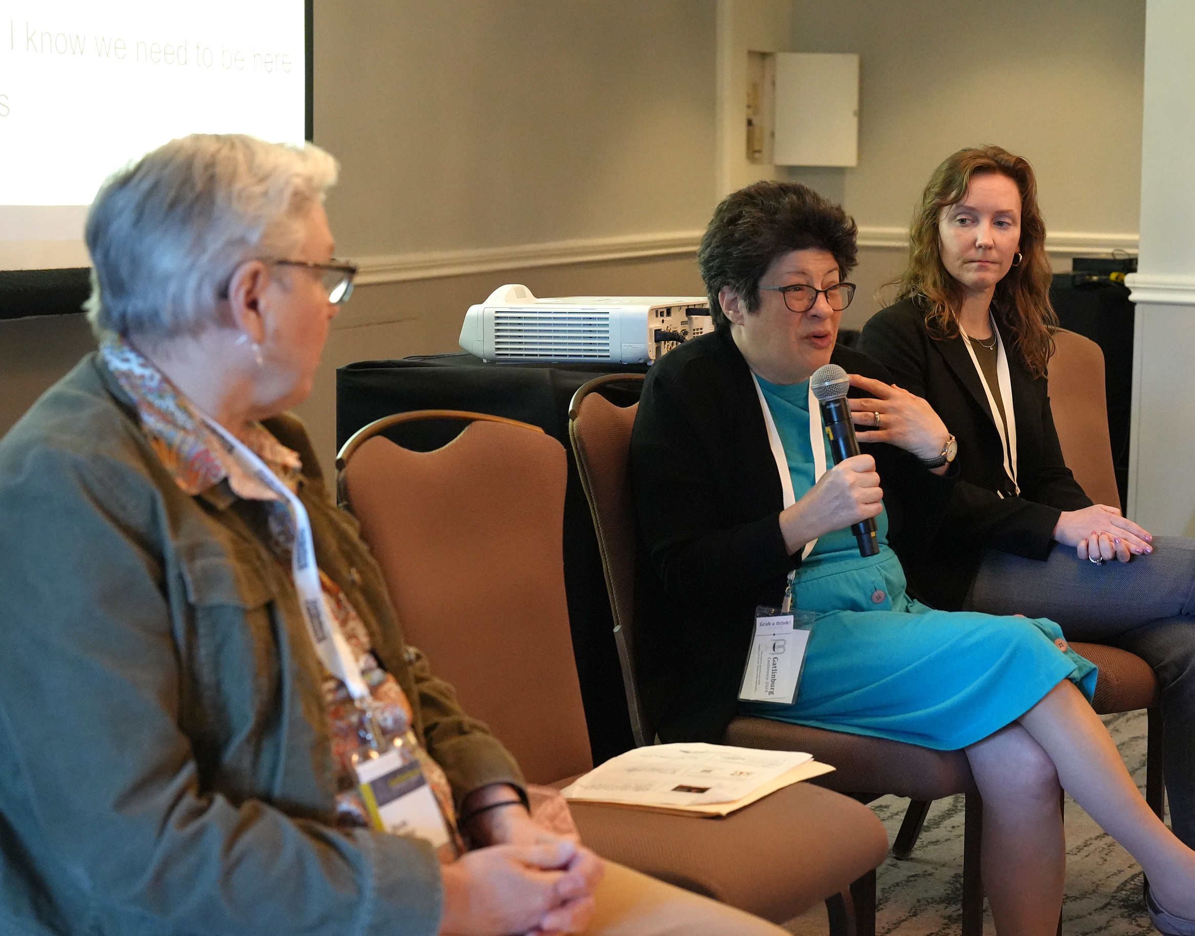 "A woman seated and holding a microphone speaks with others next to her at a panel discussion"