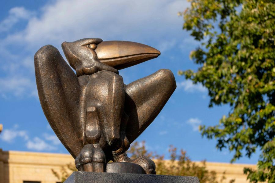 A Jayhawk statue sits in the foreground with a building and blue sky behind it.