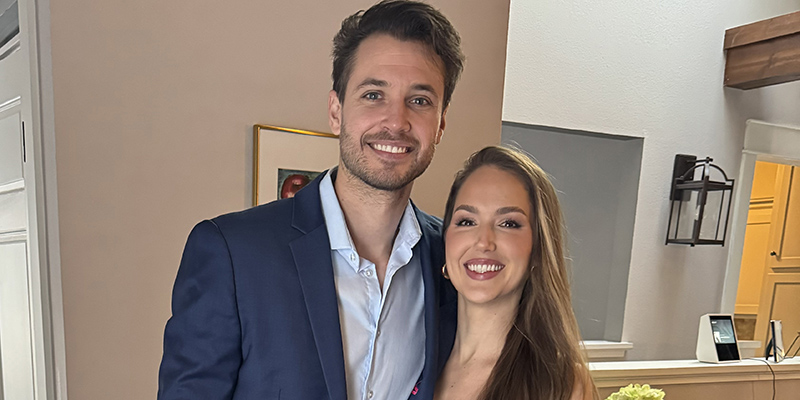 Two individuals in formal attire posing together in a room, with one wearing a blue suit and the other in a purple dress.