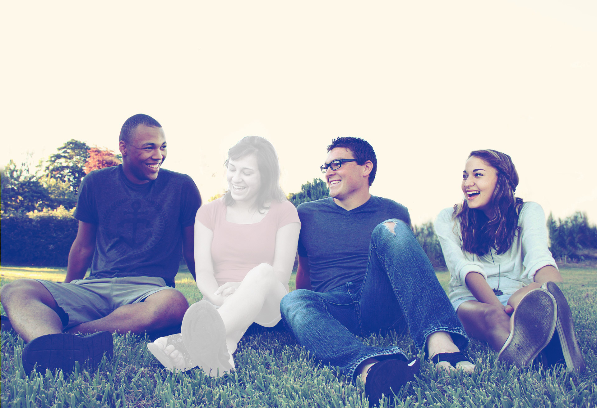 "A group of two men and two women sit in a group outside on the grassy ground, but one woman is faded while the others are clearly visible"