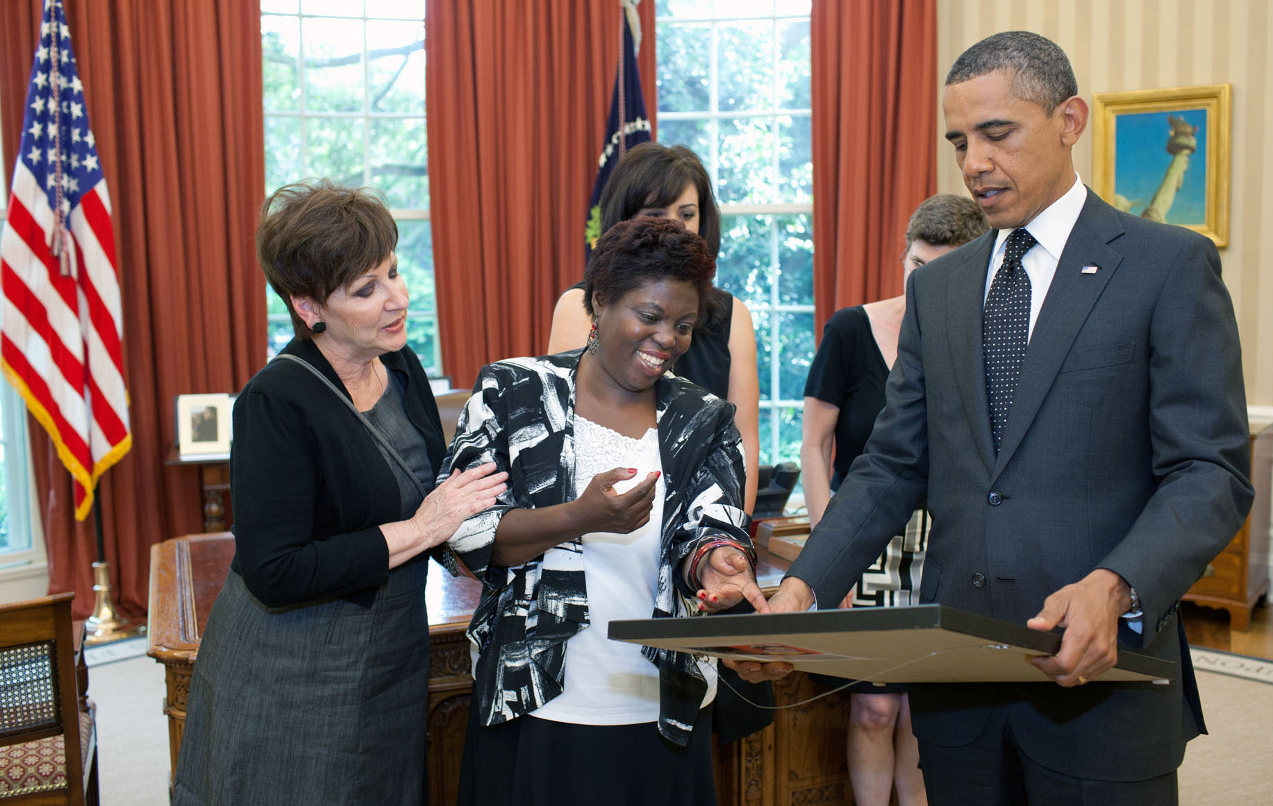 A woman with a white shirt and gray jacket presents a framed painting to President Obama in the Oval Office