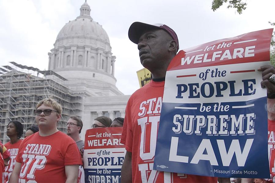 A photograph of a man holding a sign outside of the Missouri Capital. He is wearing a red T-shirt and holding signs made by Stand Up KC, an organization advocating for better pay for restaurant workers.