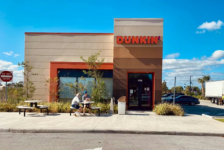 A photo of the outside of a Dunkin' sotre with people sitting at a round picnic table.
