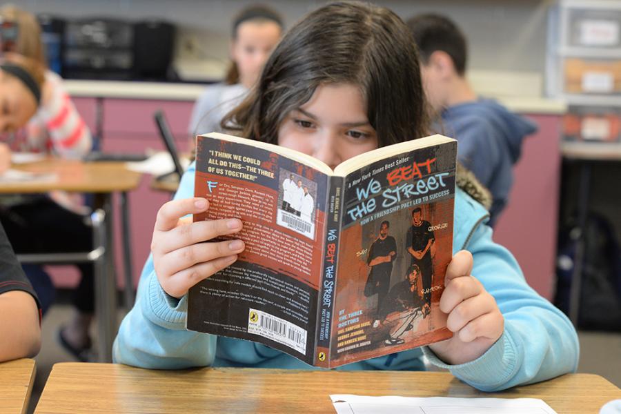 A young elementary aged girl with long brown hair is seated at a desk and intently reading a book.