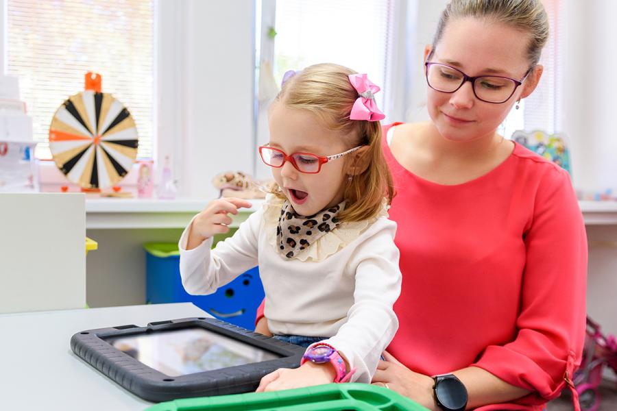 A nonverbal girl with cerebral palsy practices using a communication device while sitting on her mother's lap.