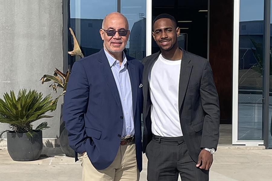 Two men wearing business jackets stand outside a modern looking building made of glass and concrete in the bakcground.