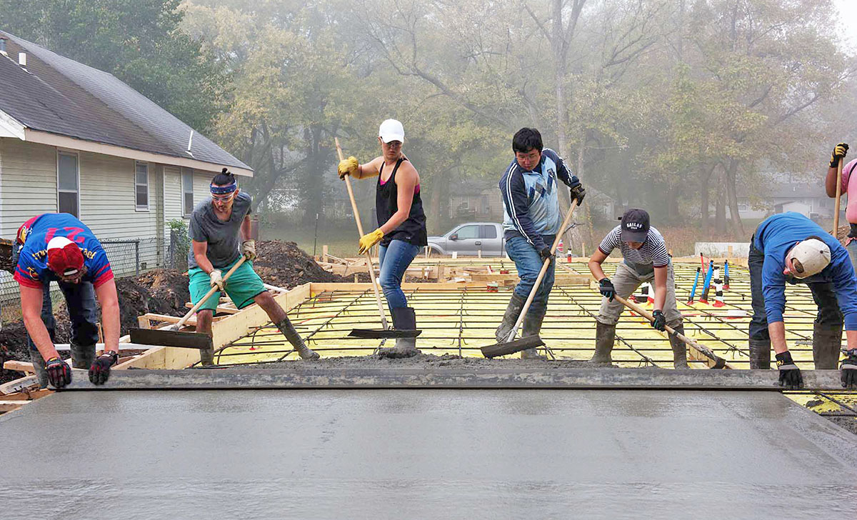 Group of students in a line, cooperatively using hand tools to smooth out a pad of concrete.